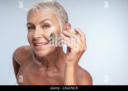 Montare e sani di età della donna in posa mentre usando il rullo di bellezza sulla sua guancia Foto Stock