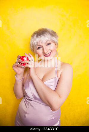 Bella curva giovane donna bionda con capelli corti gode di bere il tè da graziosi red polka dot coppa in ceramica di colore giallo su sfondo per studio. Foto Stock