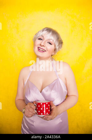 Bella curva giovane donna bionda con capelli corti gode di bere il tè da graziosi red polka dot coppa in ceramica di colore giallo su sfondo per studio. Foto Stock