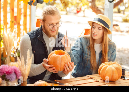 Halloween Preparaton concetto. Coppia giovane seduto a tavola all'esterno rendendo jack-o-lantern dipinto sul volto sorridente di zucca ispirato Foto Stock