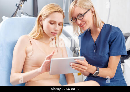 Servizio di cosmetologia. Giovane donna alla clinica di bellezza seduta la navigazione digitale compressa con la scelta del metodo close-up Foto Stock