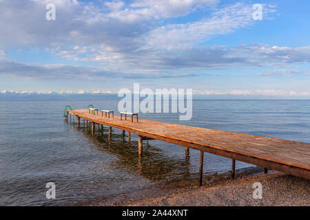 Pier con panchine sul Lago di Issyk-Kul con una catena montuosa con cime innevate all'orizzonte contro un cielo nuvoloso. Kirghizistan Foto Stock