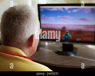 La vista laterale di un pelo grigio senior uomo seduto al tavolo e giocare con un simulatore di combattimento, essendo completamente immerso nel gioco Foto Stock