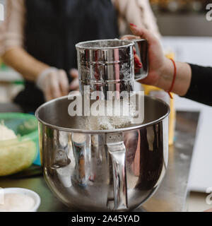 Una donna setaccia la farina in una ciotola di metallo per produrre un impasto. Le donne al master class di apprendimento sono per cuocere la pizza. Foto Stock