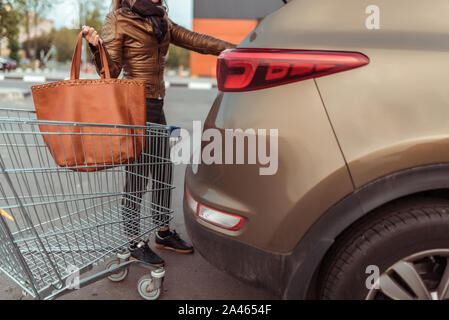 La donna nel parcheggio per automobili, aprire tronco, mette in borsa con le cose gli acquisti in auto. Carrello a ruote, estate autunno nel parcheggio al centro per lo shopping Foto Stock