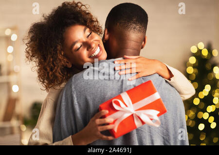 Grati afro donna con un regalo di Natale abbracciando il marito a casa Foto Stock