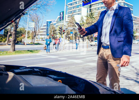 Giovane imprenditore sta parlando sul suo telefono cellulare mentre guardando sotto il cofano sollevato della sua vettura. Foto Stock