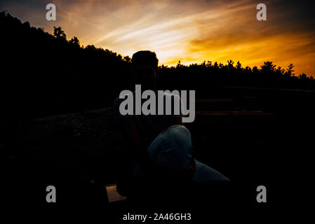Rilassata americano africano giovane uomo seduto e in attesa sulla panchina contro il cielo al tramonto Foto Stock