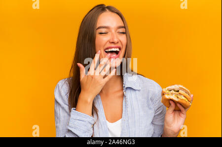 Giovane donna di mangiare hamburger e ridere in piedi su sfondo giallo Foto Stock