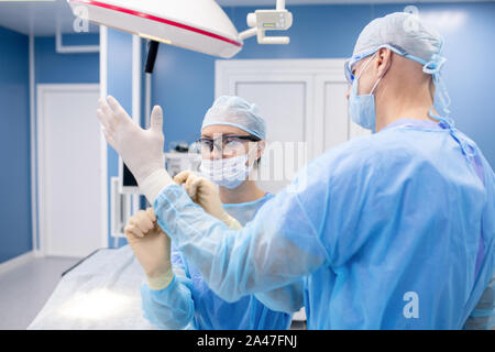 Giovane assistente in uniforme contribuendo chirurgo mettere su guanti medicali Foto Stock