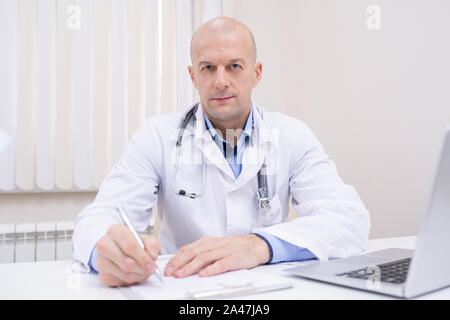 Bald di mezza età medico di fiducia in camice bianco seduta dal luogo di lavoro Foto Stock