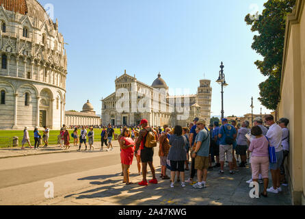 Un gruppo di turisti in appoggio all'ombra di una parete nella famosa Piazza dei Miracoli con la Torre Pendente in un caldo giorno di agosto, Pisa, Toscana, Italia Foto Stock