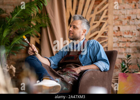 Grey-haired pensieroso pittore con il pennello seduto in poltrona in studio Foto Stock