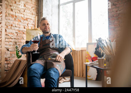 Soddisfatto artista in workwear avente un bicchiere di vino rosso mentre godendo break Foto Stock