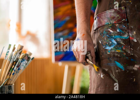 Mano dell'artista contemporaneo nel grembiule tenendo il pennello durante il lavoro in studio Foto Stock