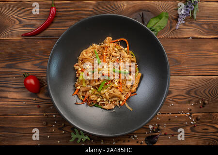 Tori udon fritto spaghetti di frumento con pollo e verdure e salsa di soia in una piastra nera su uno sfondo di legno Foto Stock