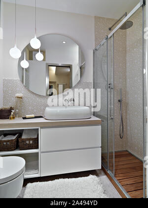 Interior shot di un moderno bagno in primo piano del lavandino armadio con un bancone lavabo e il grande specchio rotondo vicino alla cabina doccia Foto Stock