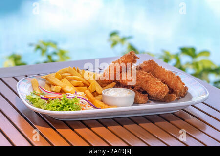 Bastoncini di pesce e patate fritte su piastra bianca con la salsa sul tavolo di legno in hotel pool bar vicino. Foto Stock