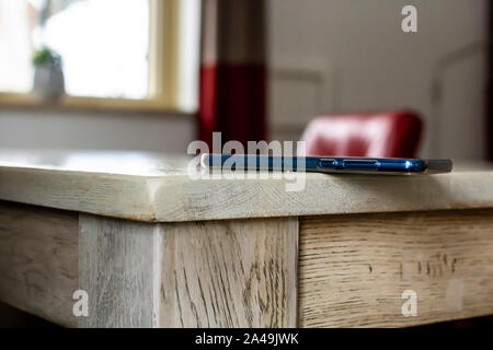 Il telefono cellulare è sdraiato pericolosamente sul bordo di un tavolo di legno, casualmente a sinistra con la possibilità di caduta, in luce naturale, contro una offuscata Foto Stock
