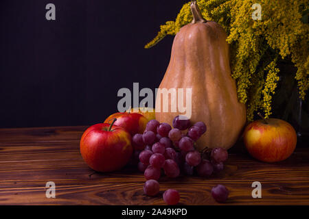 La zucca Mele Uva e fiori di colore giallo su uno sfondo nero su un tavolo di legno. Sembra una vita ancora Foto Stock