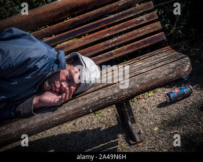 Alcolismo VAGRANT alcolica giovane uomo addormentato su una panchina nel parco in un auto indotta stupore alcolico con lattina vuota di strong lager e mozziconi di sigaretta gettati a terra nelle vicinanze. Ombre racchiudono lui come egli dorme. Foto Stock