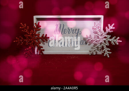 Scatola di legno con buon Natale con rosso e argento il simbolo del fiocco di neve ogni lato. sfondo rosso con luce bokeh di fondo. Prospettica piana. Foto Stock