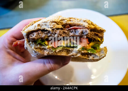 Maschio lato tenendo un morso, deliziosa cheeseburger, sullo sfondo di una piastra. Close-up. Foto Stock