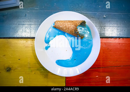 Blue gelato fuso e fatto fluire al di fuori del cono di cialda su una piastra bianca che si erge su un multi-colore tabella. Close-up. Foto Stock