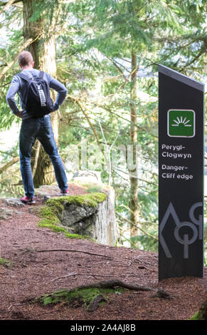 Uomo che guarda sulla cima di una scogliera con un cartello di avvertimento in gallese e inglese "ira Cliff Edge". Foto Stock