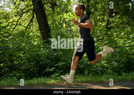 Giovani runner africani la formazione nella foresta nella mattina di sole. Sportivo maratona in esecuzione sulla passerella del parco e ascoltare musica. Atleta che indossa in bianco sneakers e calze, nero di t-shirt e shorts. Foto Stock