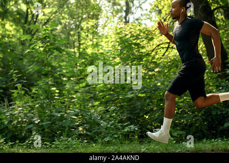 Muscolare, la via e il campo atleta che corre maratona in foresta nella mattina di sole. Giovane africano corpo sportivo ascoltando musica con le cuffie. Concetto di salute e di sport. Foto Stock