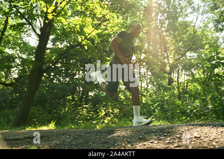 Bello atleta che corre nel parco la passerella nella mattina di sole. African atleta indossa in bianco sneakers, nero di t-shirt e shorts. Una forte formazione di runner nella foresta. Foto Stock