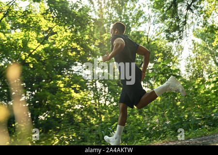 African sportivo a correre e ad ascoltare musica con le cuffie. Atleta che indossa in nero di t-shirt e shorts, bianco sneakers e calzini formazione nel parco in mattina d'estate. Foto Stock