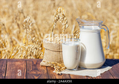 Vegano Latte di avena in vetro e la caraffa con il sacco di chicchi sulla tabella sopra contro mature campo di cereali Foto Stock