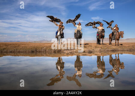 Ritratto di gruppo di quattro tradizionali kazake cacciatori di eagle con loro le aquile reali riflettendo in acqua di fiume. Ulgii, Mongolia occidentale. Foto Stock