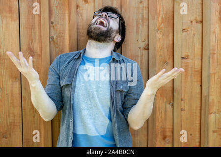 Un uomo in preda alla disperazione alza le palme e della testa e grida a voce alta. Espressione delle emozioni dei problemi. Isolato sullo sfondo di un vecchio muro in legno. Chiudere Foto Stock