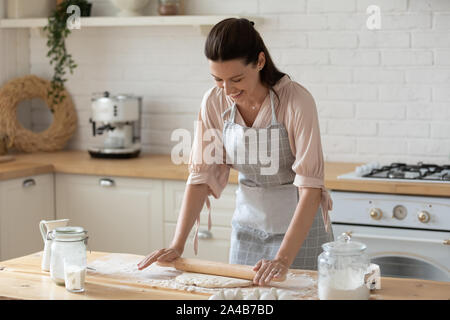 Sorridente giovane donna che indossa il grembiule di stendere la pasta in cucina Foto Stock