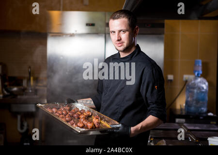 Ritratto chef maschio con cibi cotti in piedi in cucina. Cucina a tema. giovane uomo caucasico in nero uniforme, guanti in lattice al ristorante in cucina Foto Stock