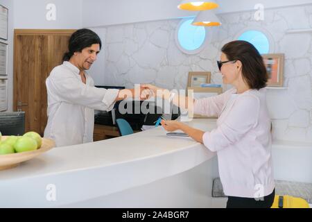 Controllo degli ospiti in hotel, receptionist maschio in piedi presso la reception a parlare e saluto donna valutazione Foto Stock