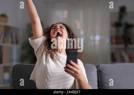 Excited donna celebra il successo tenendo il telefono cellulare seduto su un divano nella notte a casa Foto Stock