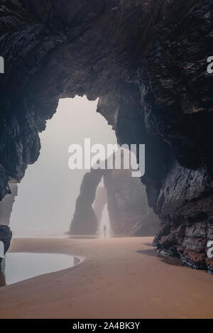 Silhouette di un giovane uomo nella nebbia tra archi in Playa las Catedrales. Cattedrali beach a Ribadeo Galizia di Lugo Spagna Foto Stock