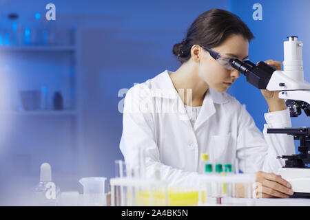 Vista laterale Ritratto di giovane donna scienziato cercando nel microscopio, mentre facendo ricerca in laboratorio medico illuminato da luce blu, spazio di copia Foto Stock