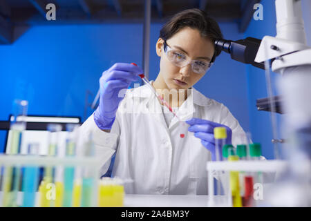 Angolo basso Ritratto di giovane donna scienziato preparazione campione di sangue usando un contagocce mentre si lavora sulla ricerca medica in laboratorio Foto Stock