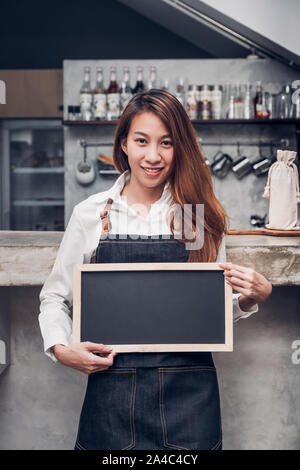 Giovani asiatici barista indossare jean grembiule azienda lavagna vuota nel menu di caffè al banco bar con sorriso faccia,servizio di caffetteria concetto,titolare di azienda start upla Foto Stock
