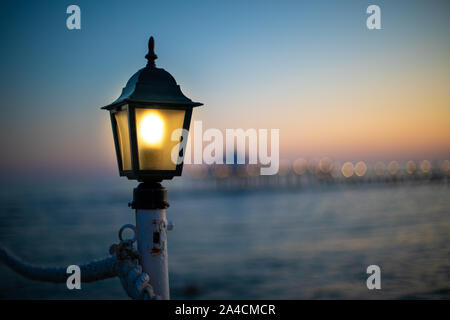 Una lanterna su un molo si illumina di colore giallo brillante al crepuscolo Foto Stock