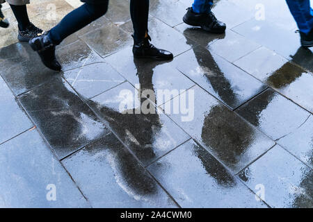 Sfocate gambe di riflessione e le ombre e sagome delle persone in condizioni di bagnato il marciapiede, in una pozza di persone a piedi su un sensore pioggia Foto Stock