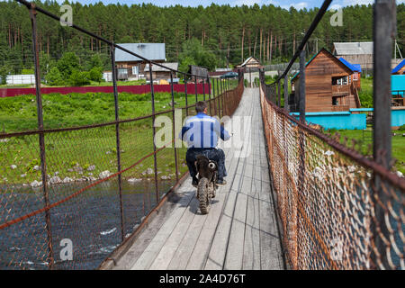 Un motociclista giostre di un motociclo fuoristrada in sospeso un ponte di legno su un fiume di montagna. Foto Stock