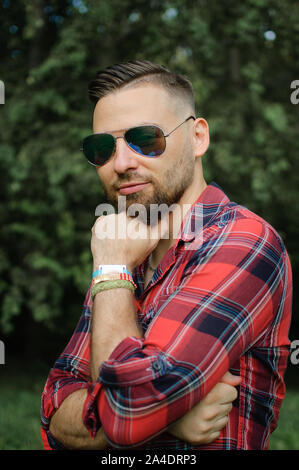 All'esterno Ritratto di giovane uomo barbuto con occhiali da sole guardando la telecamera nel parco su alberi sullo sfondo Foto Stock