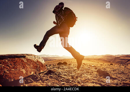 Escursionista uomo salta con grande zaino per un rock contro le montagne e il sole al tramonto Foto Stock