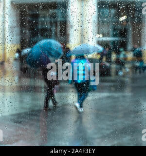 Astratti persone sotto l'ombrello, tempo piovoso, offuscata motion. Vista attraverso il vetro della finestra con gocce di pioggia sul vetro con street bokeh luci Foto Stock